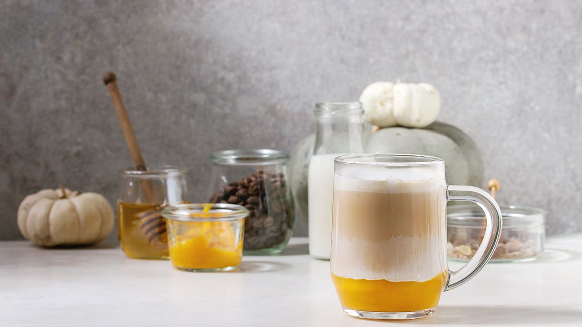 Glass of pumpkin layered spice latte with pumpkin puree, with milk foam and cinnamon standing with ingredients in jars and decorative white pumpkins on white marble table with gray wall at background.