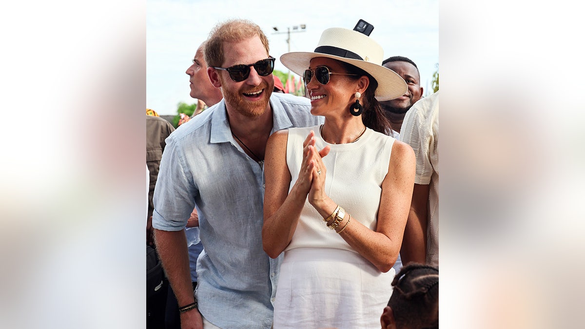 Prince Harry laughs in a blue t-shirt and sunglasses on with Meghan Markle in a cream top and hat and white skirt