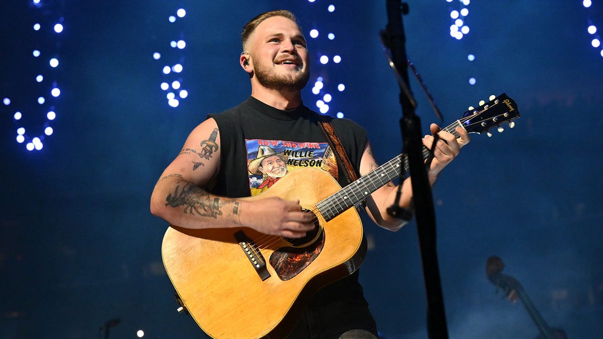 Zach Bryan looks up in a black cut-off shirt smiling playing the guitar on stage