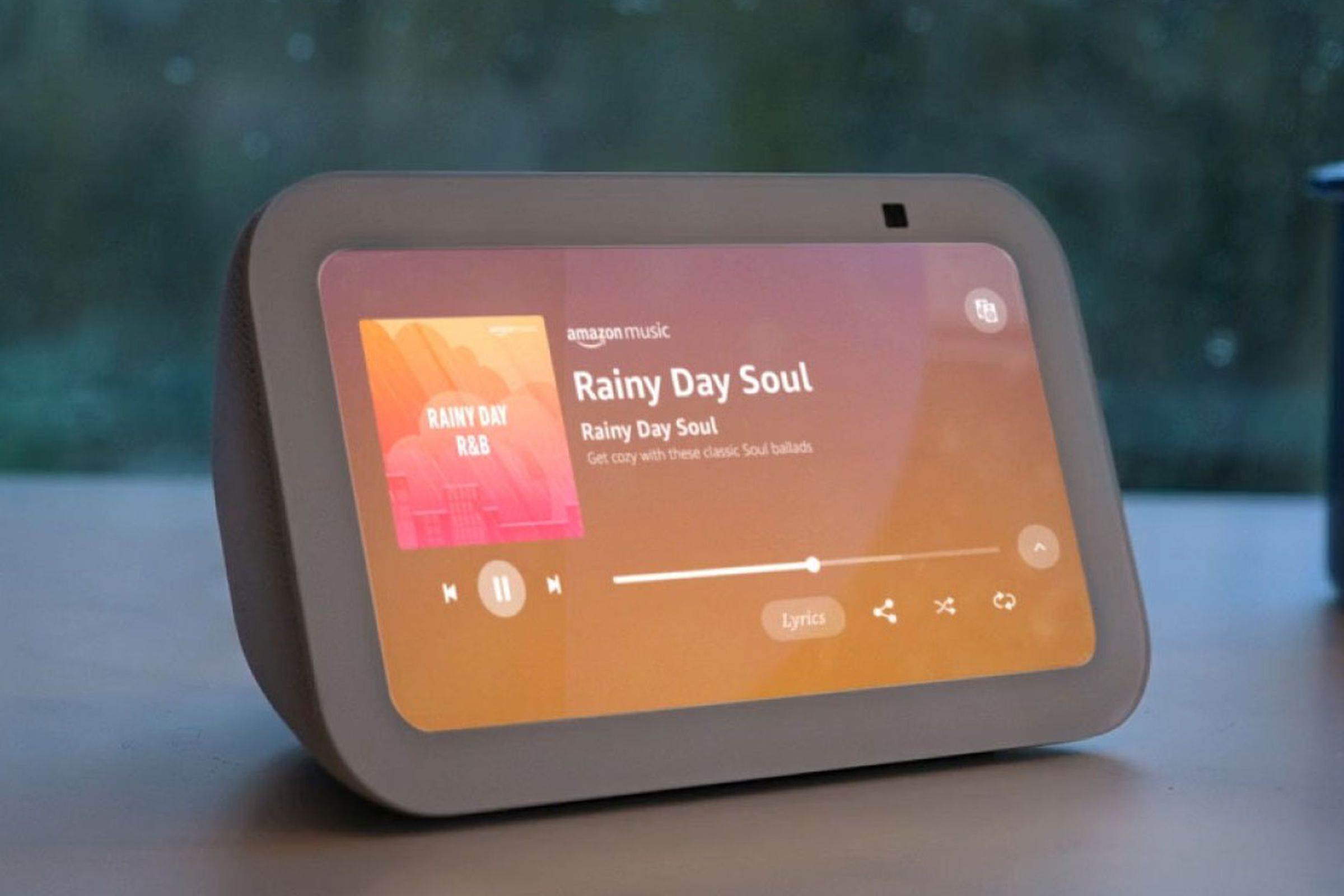 The third-gen Echo Show 5 turned on while on a desk in front of the window during a rainy day.