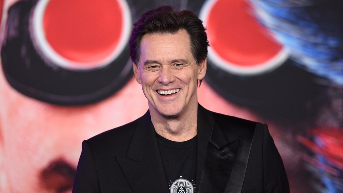 Jim Carrey in a black shirt and black jacket laughs on the carpet in Hollywood