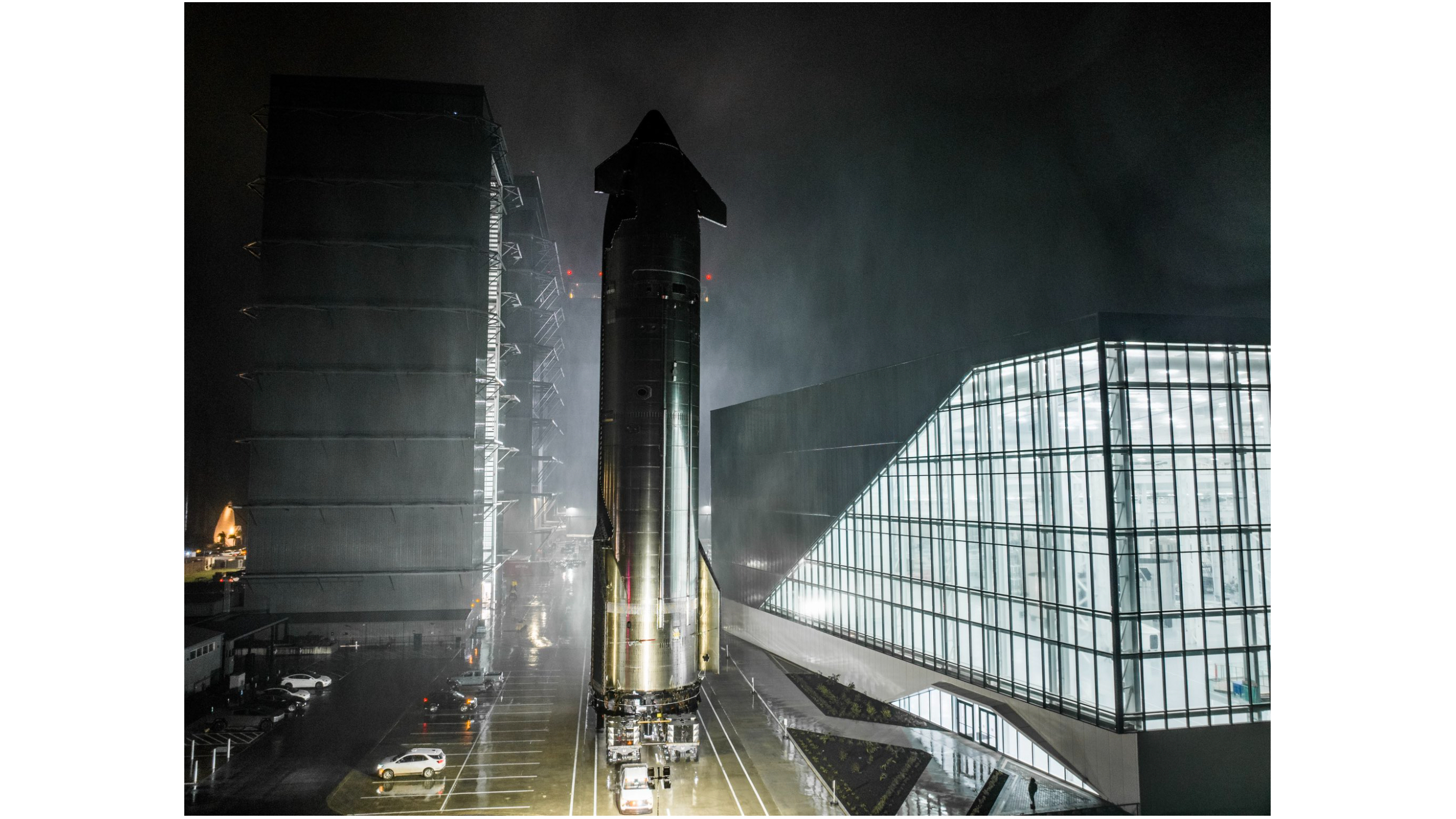 a large silver rocket rolls down a road at night, passing close to a lit-up building