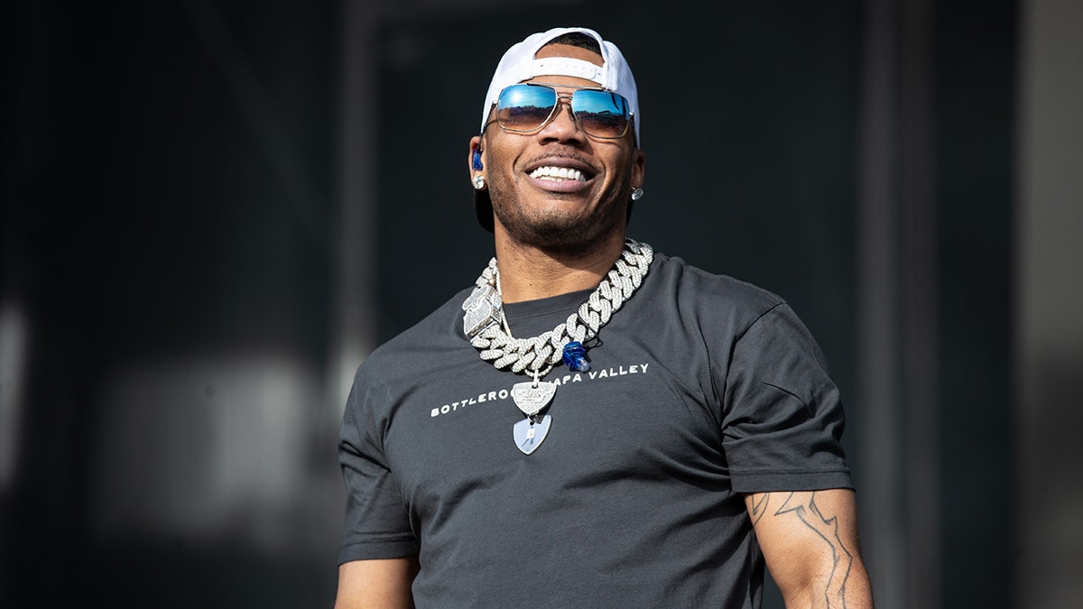 Nelly looks up on stage wearing a black t-shirt, a huge silver chain, backwards hat, and blue-tinted sunglasses