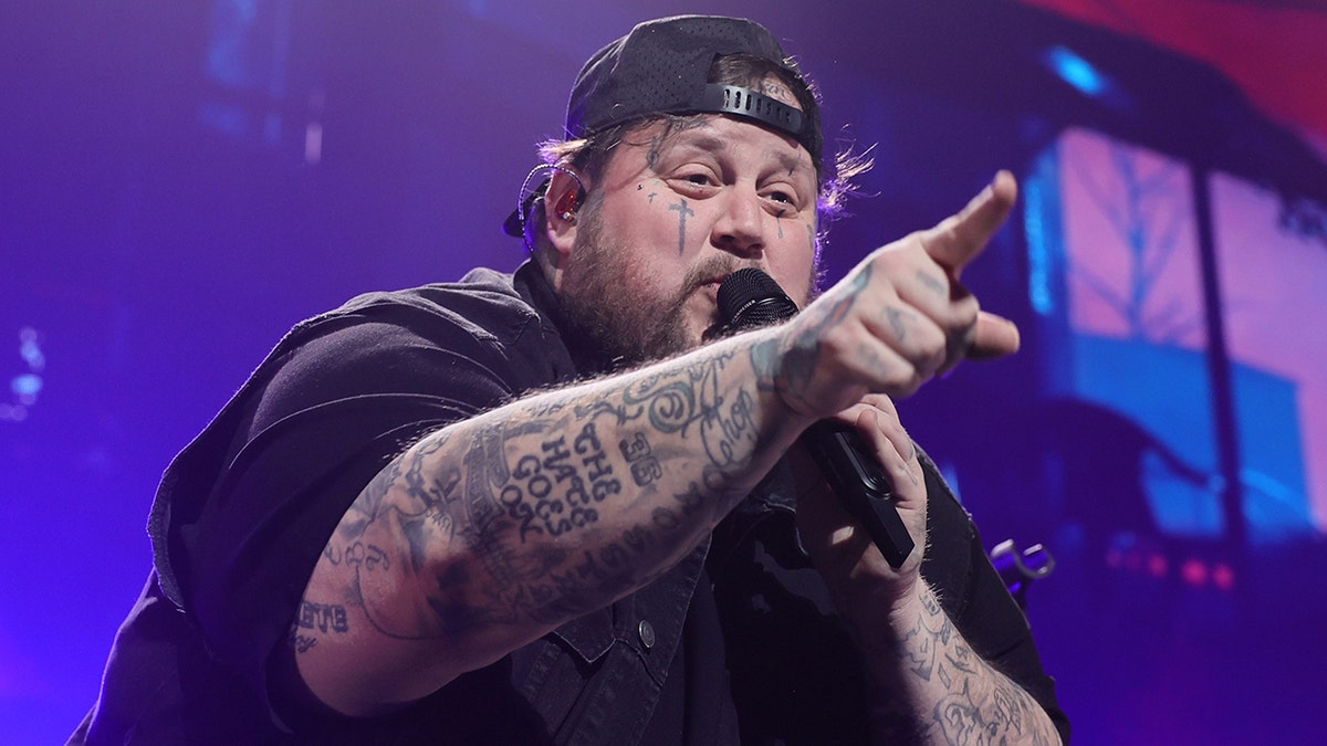 Jelly Roll wearing a backwards black hat and black shirt points at the audience while performing