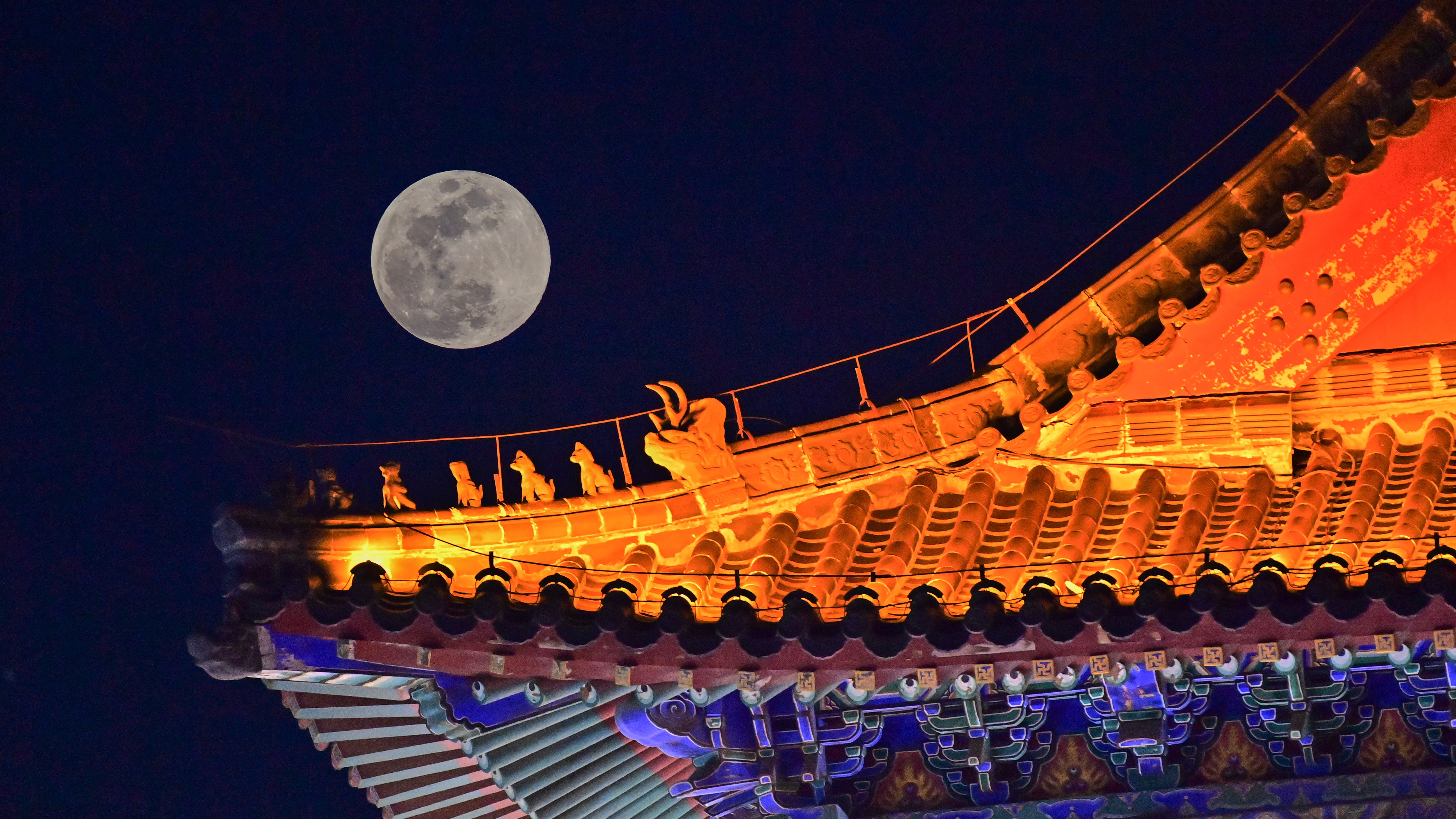 full moon on the left shining next to an ornate roof of a building illuminated by lights.