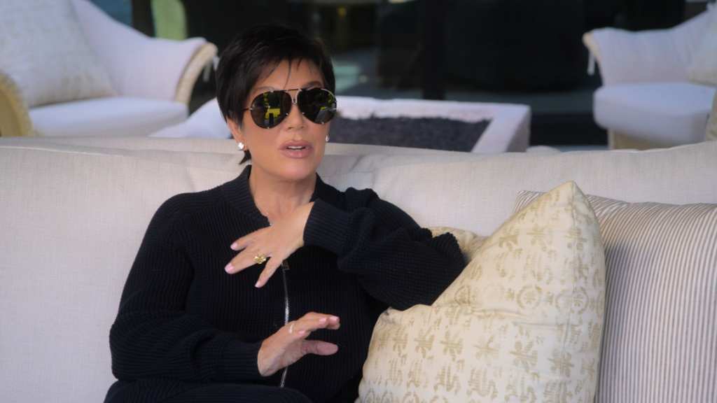 Kris Jenner wears shades and sits on oversized furniture.