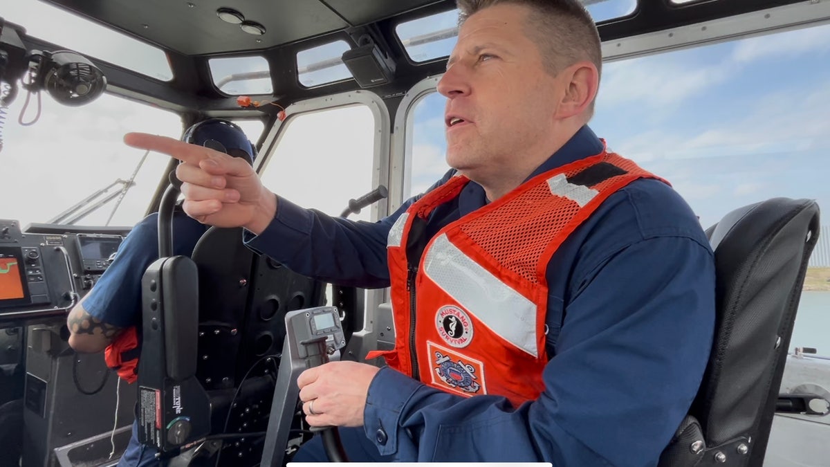 A U.S. Coast Guard Captain wearing a neon orange life jacket points to debris belonging to suspected smugglers along the Rio Grande