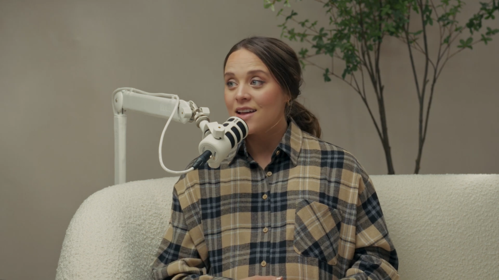 Jinger Duggar speaks into a mic while wearing a beige plaid shirt.