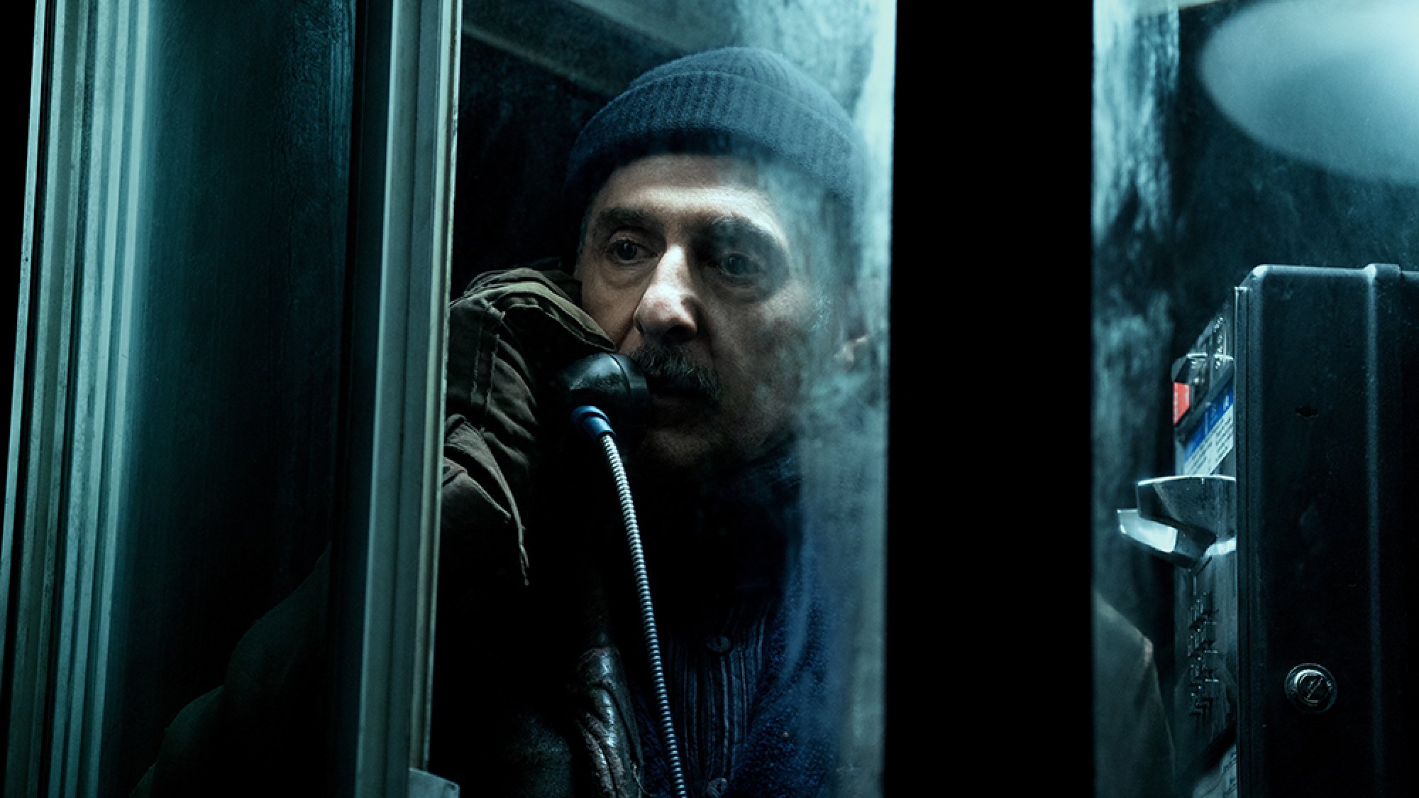 A man in a dark hat stands talking on a payphone at night.