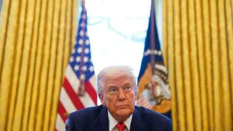 Donald Trump is seated at a desk in the Oval Office